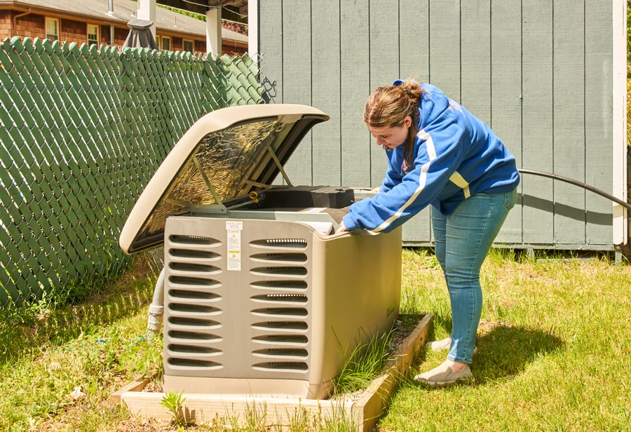 A woman in a LaPlante blue jacket and jeans opens an outdoor generator to inspect it beside a green fence and gray shed on a grassy lawn.