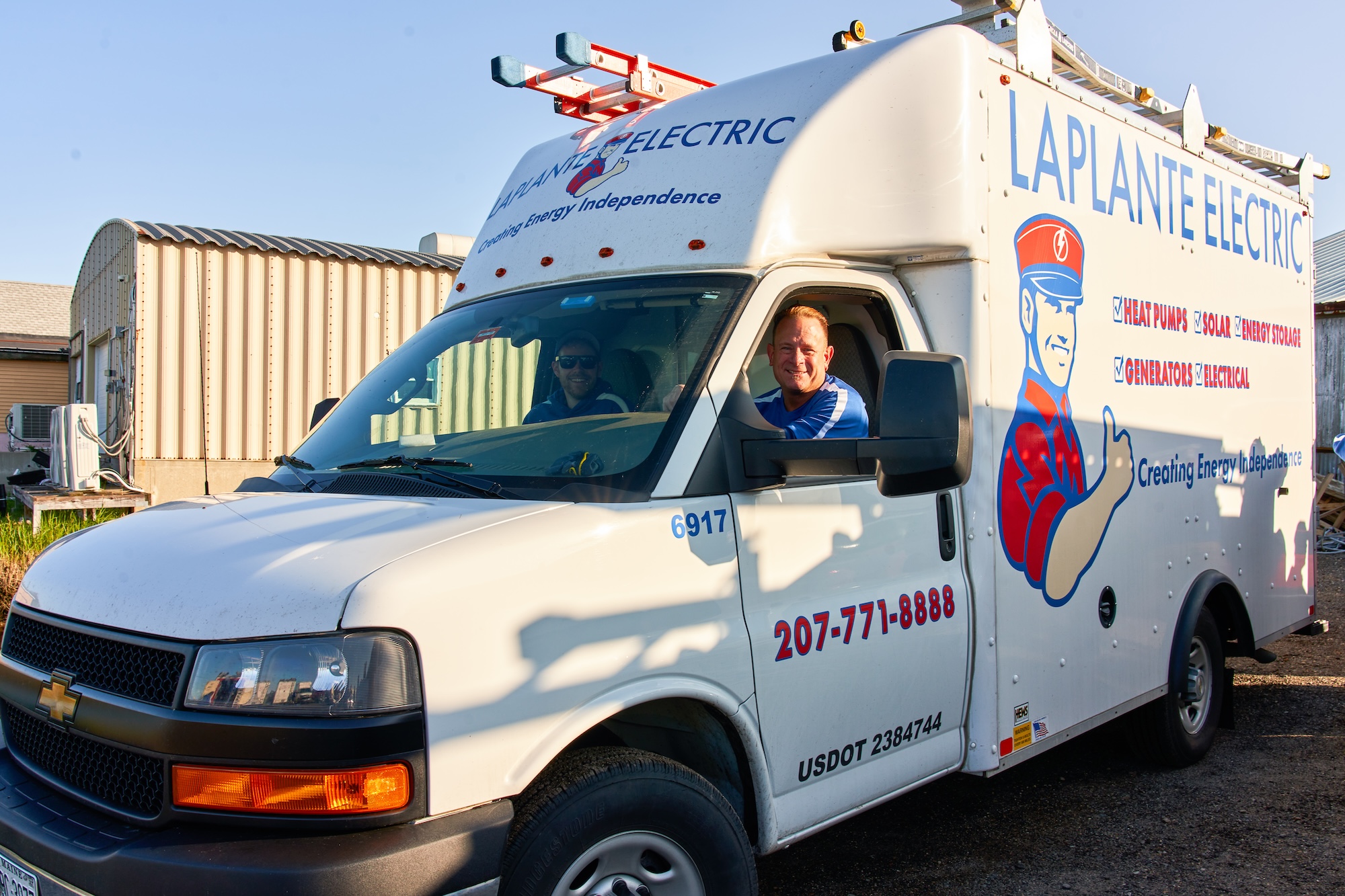 A white LaPlante Electric service truck, with a logo and contact information on the side, parks against a backdrop of industrial buildings. Two men smile through the window, representing the right choice for your electrical needs. Equipped with a ladder on top, the truck stands ready to install your home's new heat pump water heater.