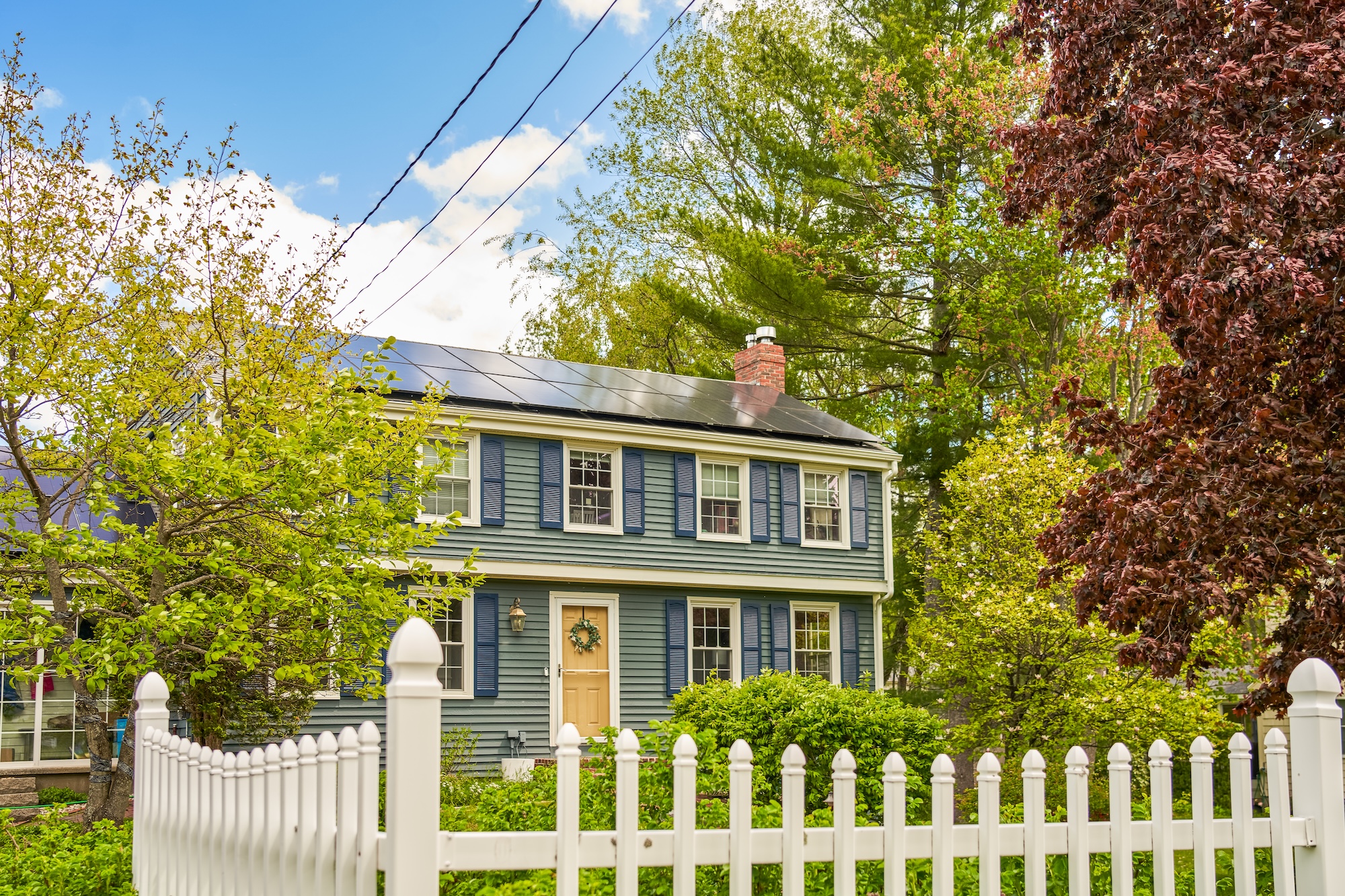 A charming two-story house with blue siding and a beige door showcases black shutters and solar panels, making it solar ready. Lush greenery and trees surround the house, while a white picket fence stands in front. The scene unfolds under a partly cloudy sky.