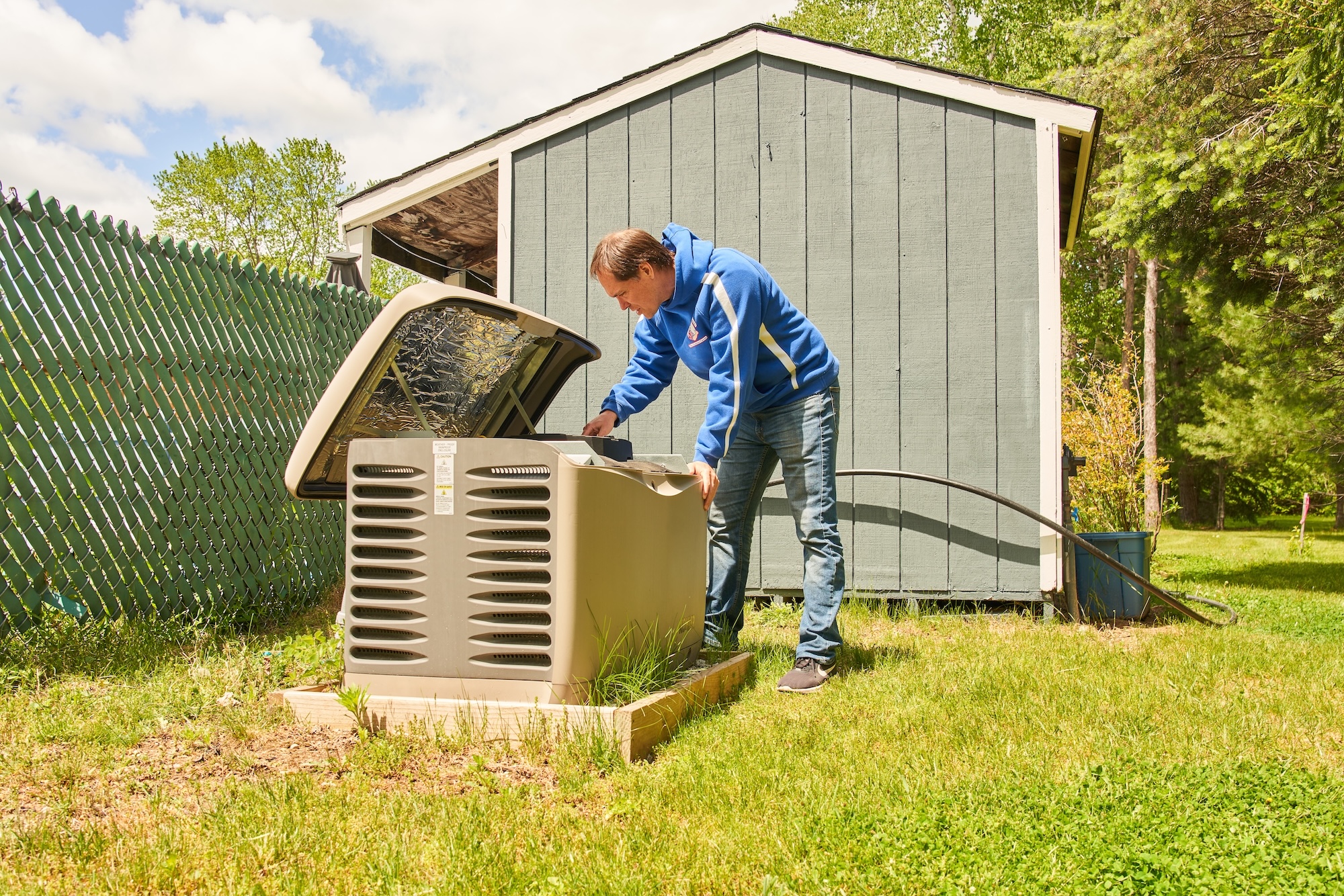 A man in a blue hoodie regularly maintains a reliable generator with its lid open, next to a gray shed on the grassy lawn. A green fenced area and trees stand in the background under a partly cloudy sky.