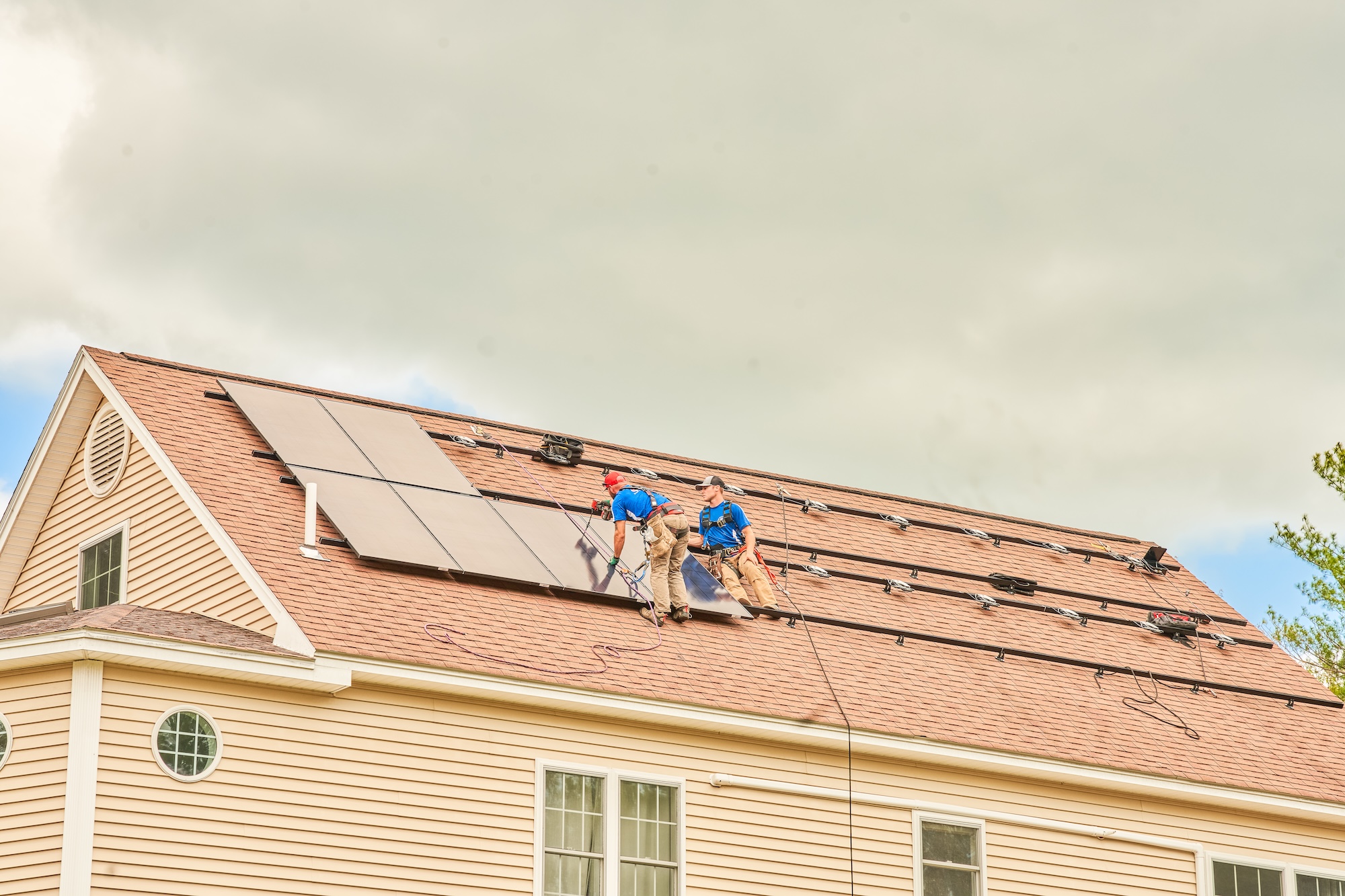 Two workers in blue shirts install solar panels on the sloped roof of a beige house under a cloudy sky, following home safety measures as they work. They have already secured some panels and are mounting the others.