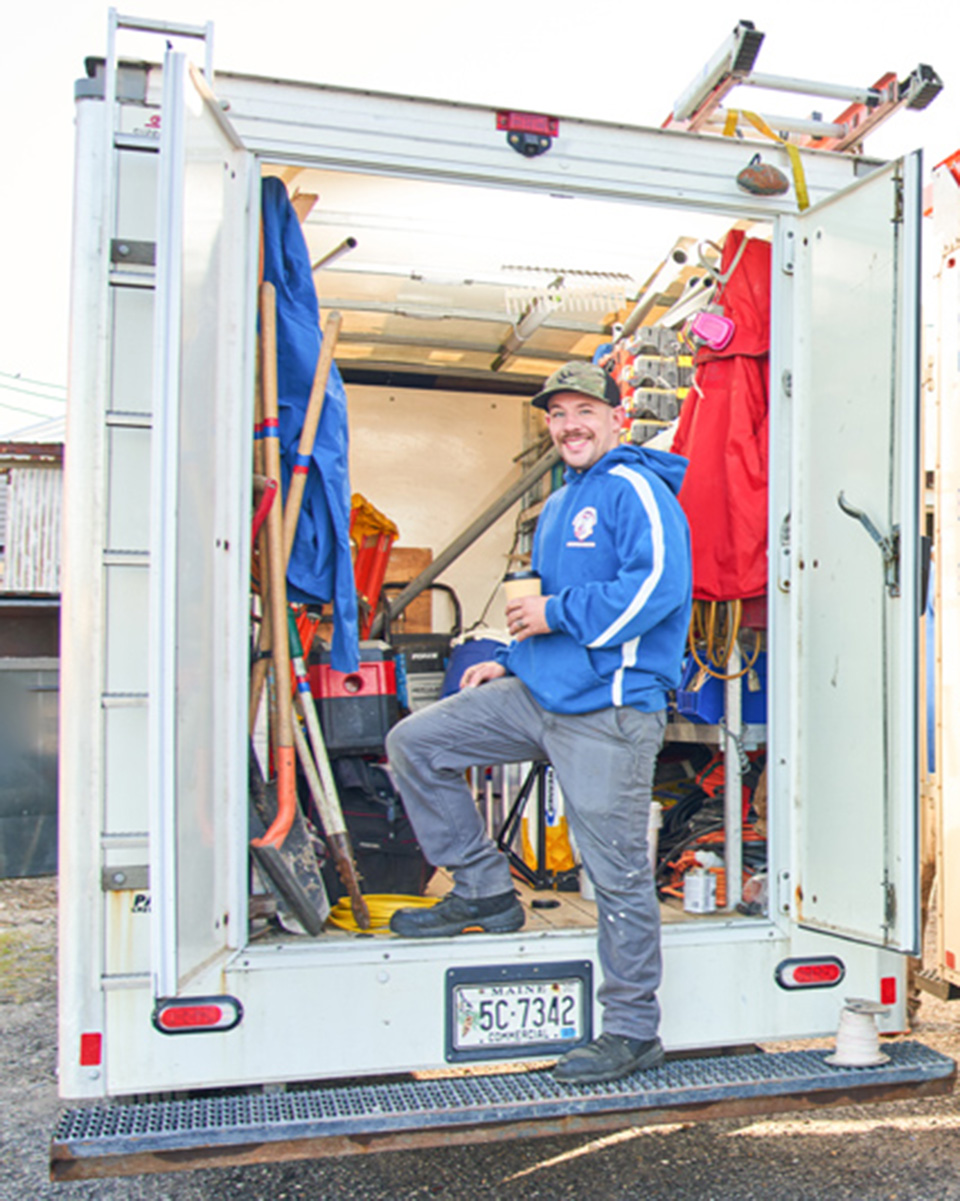 A smiling worker in a blue hoodie stands in the back of a work van, holding a cup and looking at the camera. Plumbing repair tools and equipment fill the van, and with the doors open, you can see various tools and supplies inside.