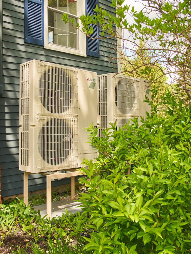 Two outdoor heat pump units sit on a stand beside a blue Maine house with white trim and shutters, surrounded by green bushes.