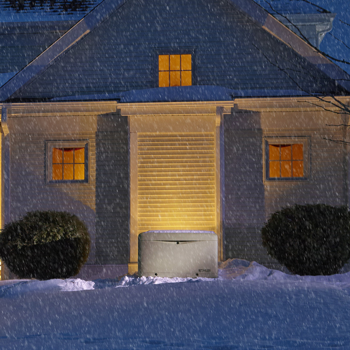 Snow falls at night as yellow lights glow from the house windows. An electrical generator sits by the garage amid snowy bushes.
