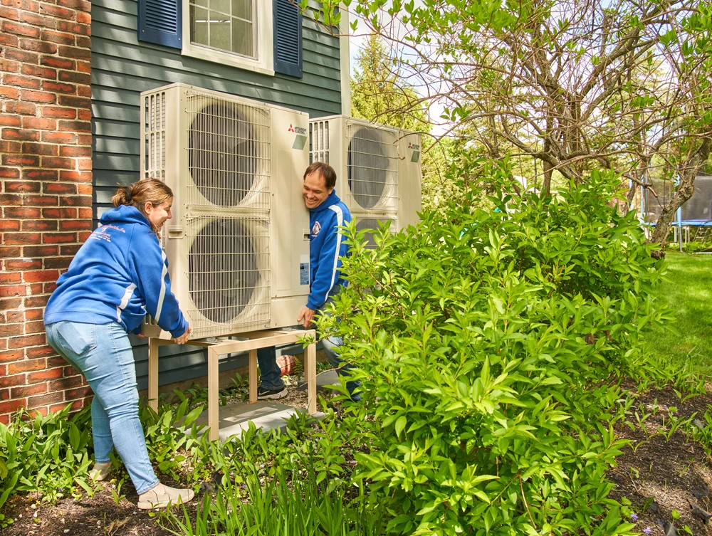 Two people in blue hoodies install large outdoor heat pump units on a metal stand beside a house in Scarborough, ME, with green bushes.