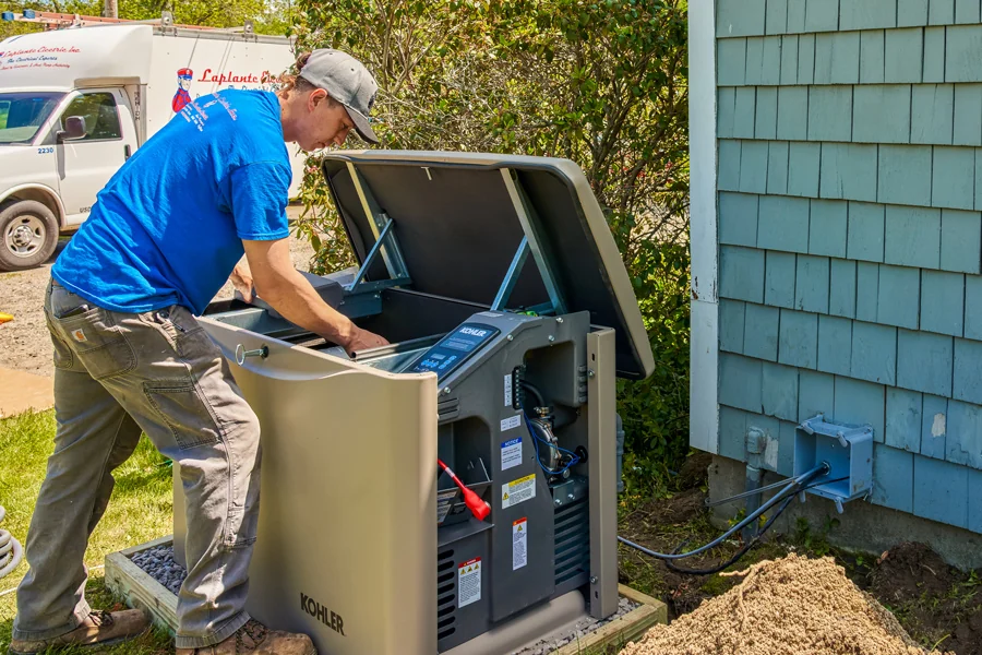 An electrician in a blue shirt repairs an open standby generator by a Maine house, with tools inside and cables linked to the building.