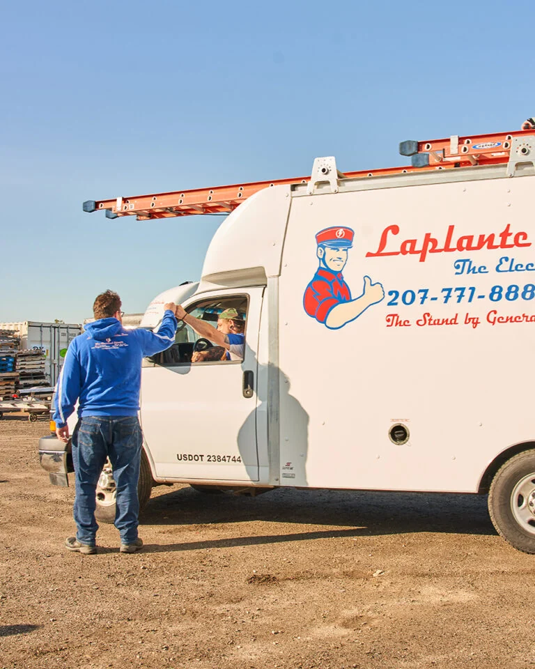 A LaPlante electrician outside high-fives a Maine electrician in a white utility van labeled "Laplante," with ladders and a cartoon logo on top.