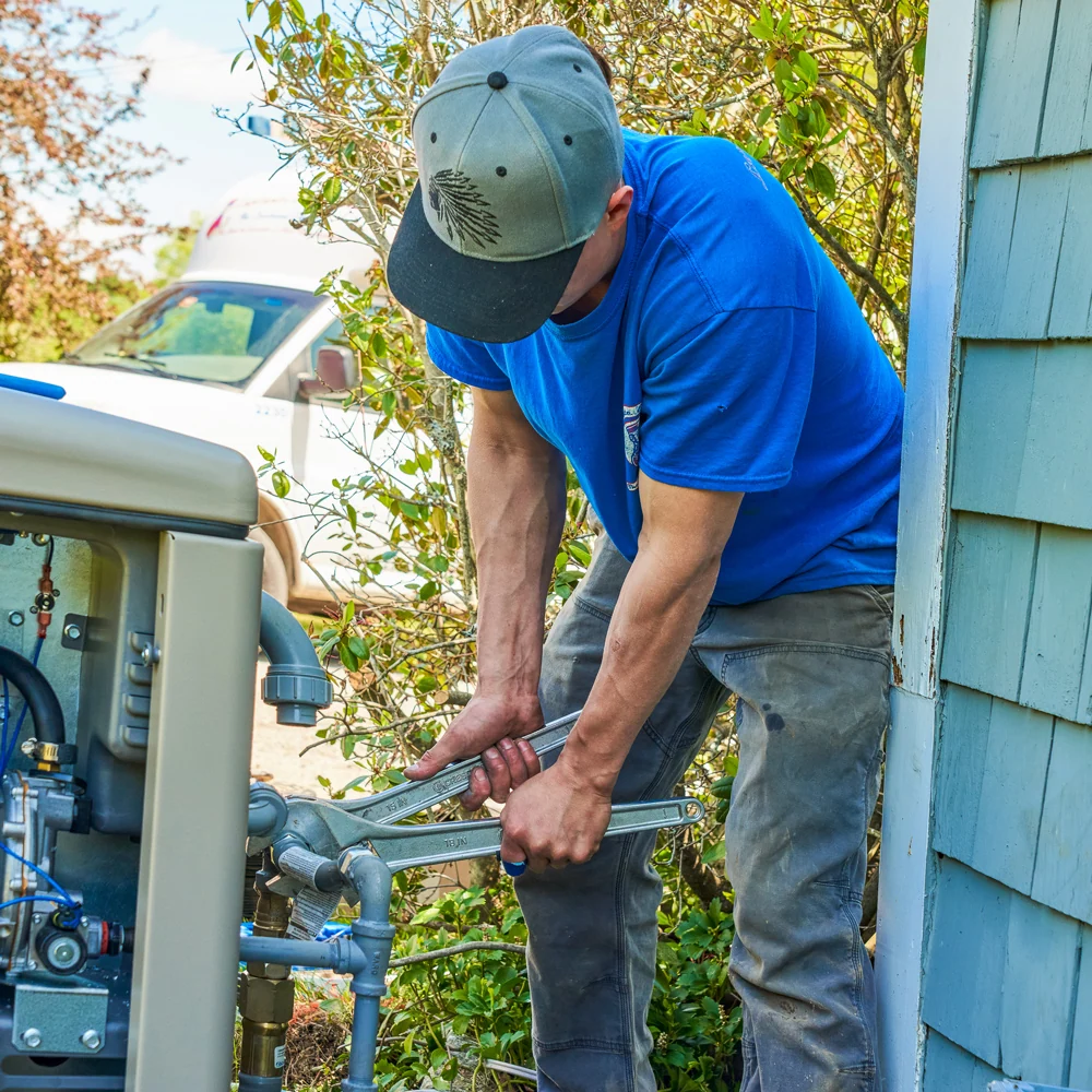 A LaPlante electrician in a blue shirt and gray cap works on outdoor piping with a large wrench near a building in Scarborough, ME; bushes and a white vehicle sit in the background.