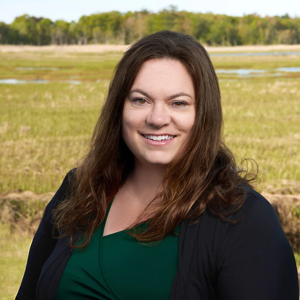 A woman with long brown hair and a green top stands outdoors near Scarborough ME, smiling in front of grass, trees, and blue sky.